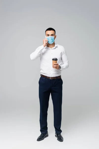 Full length view of hispanic businessman with coffee to go talking on mobile phone on grey — Stock Photo