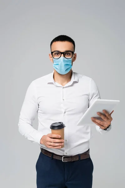 Joven empresario hispano en máscara médica sosteniendo tableta digital y café para ir aislado en gris - foto de stock