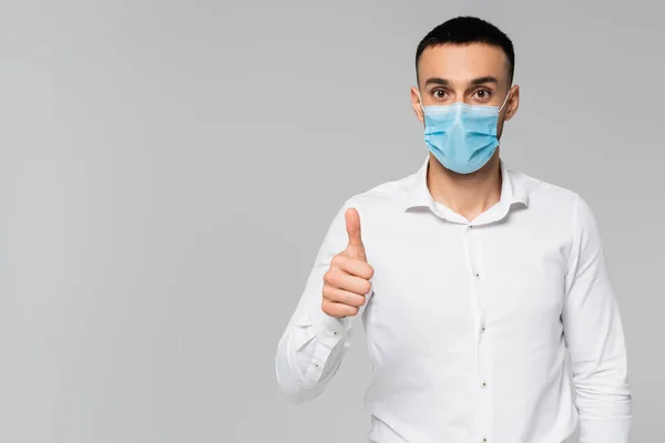 Morena hispano hombre de negocios en máscara médica mostrando el pulgar hacia arriba aislado en gris - foto de stock