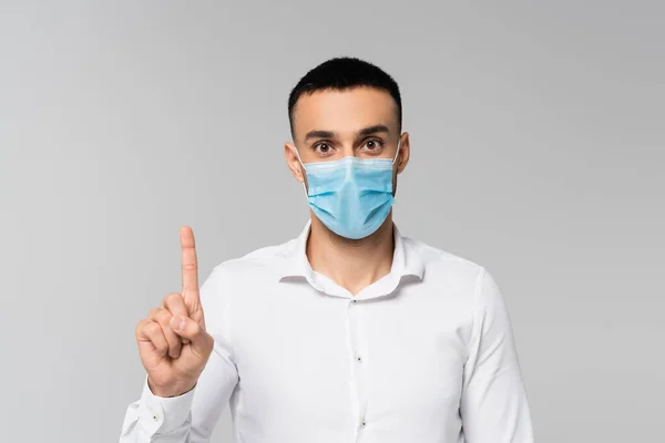 Jeune gestionnaire hispanique en masque médical pointant vers le haut avec le doigt isolé sur gris — Photo de stock