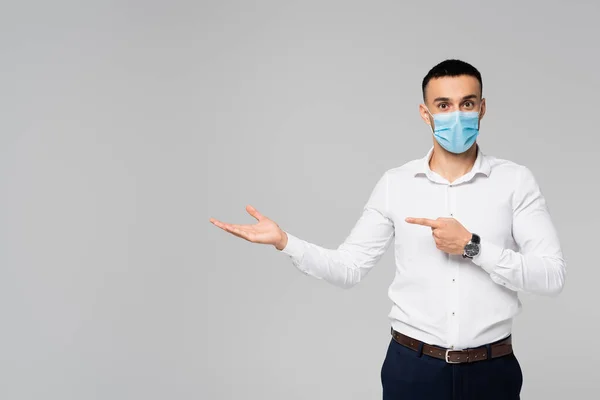 Empresario hispano en máscara médica señalando a un lado mientras mira la cámara aislada en gris - foto de stock