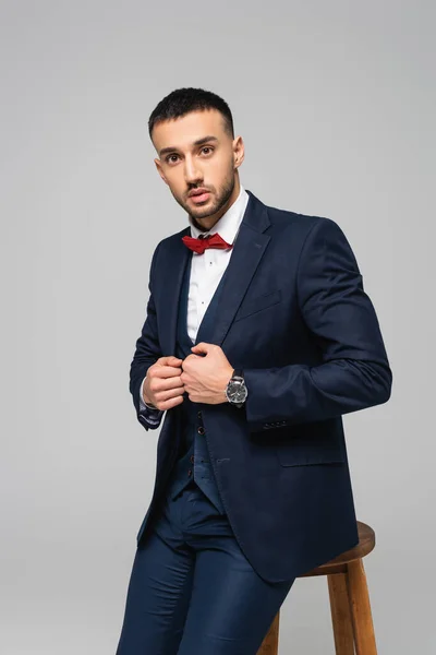 Elegant hispanic man in blue suit looking at camera while posing near high stool isolated on grey — Stock Photo