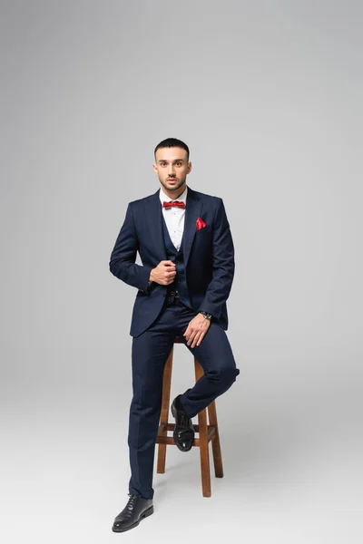 Full length view of young hispanic man in blue suit posing near high stool on grey — Stock Photo