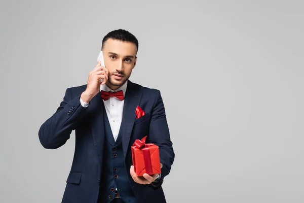 Morena hombre hispano en traje elegante hablando en el teléfono inteligente mientras sostiene caja de regalo roja aislada en gris - foto de stock