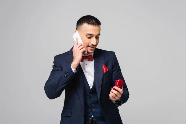 Sonriente hombre hispano en traje azul hablando en teléfono inteligente mientras sostiene joyero aislado en gris - foto de stock