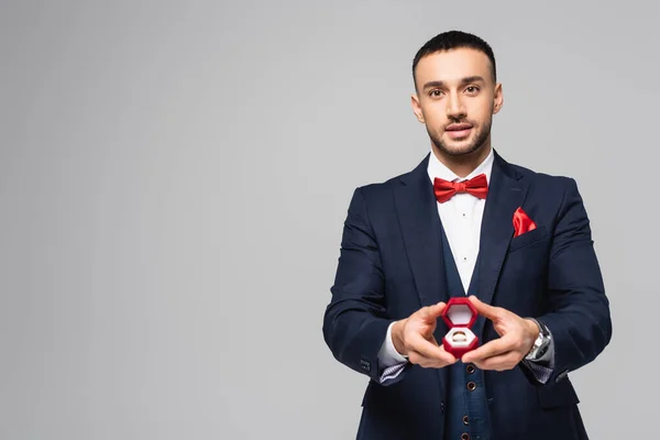 Young hispanic man in elegant suit holding red jewelry box with wedding ring isolated on grey — Stock Photo