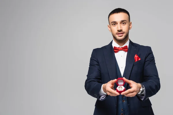 Jeune homme hispanique en costume élégant tenant boîte à bijoux rouge avec anneau de mariage isolé sur gris — Photo de stock