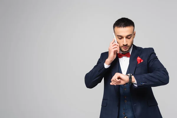 Елегантний іспаномовний чоловік перевіряє час на наручному годиннику під час розмови на смартфоні ізольовано на сірому — стокове фото