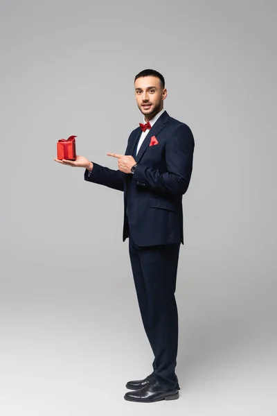 Vista completa del elegante hombre hispano señalando con el dedo a la caja de regalo roja en gris - foto de stock