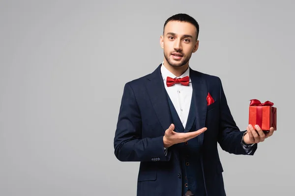 Young hispanic man in elegant suit pointing with hand at red gift box isolated on grey — Stock Photo
