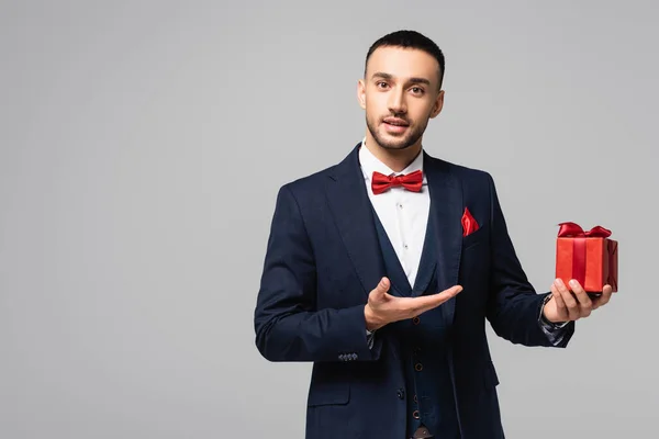 Joven hombre hispano en traje elegante apuntando con la mano a la caja de regalo roja aislada en gris - foto de stock