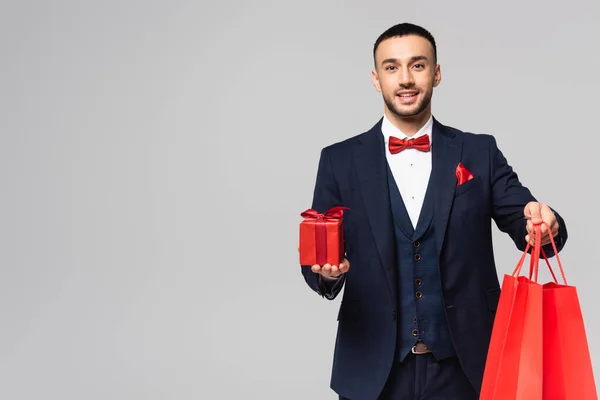 Elegante hombre hispano de traje azul con caja de regalo roja y bolsas aisladas en gris - foto de stock