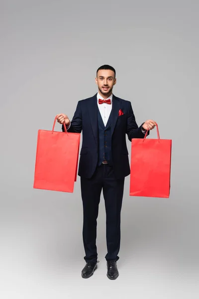 Vue pleine longueur de l'homme hispanique heureux en costume élégant tenant sacs à provisions rouges sur gris — Photo de stock