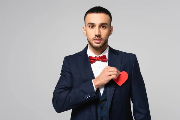 Elegante hombre hispano mirando a la cámara mientras sostiene el corazón cortado de papel rojo aislado en gris - foto de stock