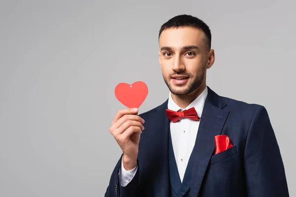 Élégant homme hispanique souriant à la caméra tout en tenant rouge coeur coupé en papier isolé sur gris — Photo de stock