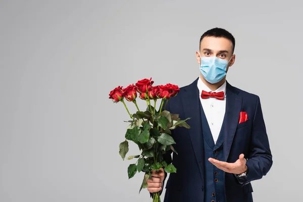 Joven hispano de traje azul y máscara médica apuntando a rosas rojas aisladas en gris - foto de stock