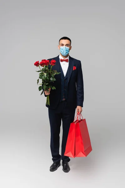 Ganzkörperansicht des eleganten hispanischen Mannes in medizinischer Maske mit roten Rosen und Einkaufstaschen auf grau — Stockfoto