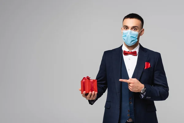 Jeune homme hispanique en costume élégant et masque médical pointant vers boîte cadeau rouge isolé sur gris — Photo de stock