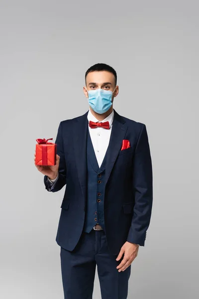 Elegante uomo ispanico in maschera medica mostrando scatola regalo rossa isolata su grigio — Foto stock