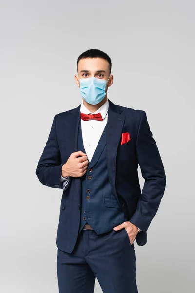Элегантный латиноамериканец в медицинской маске, стоящий с рукой в кармане, изолированный на сером — стоковое фото