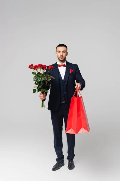 Vista completa del elegante hombre hispano sosteniendo bolsas de compras rojas y rosas rojas en gris - foto de stock