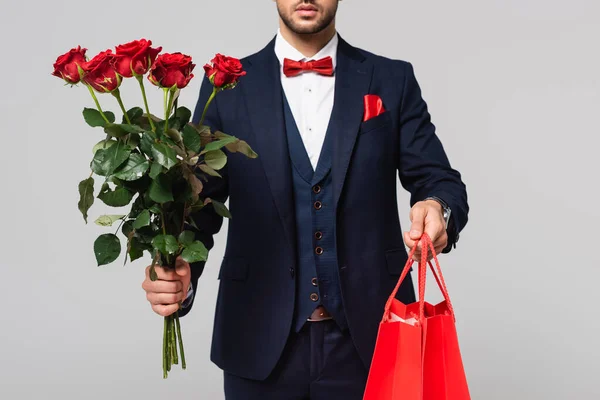 Обрезанный вид человека в элегантном костюме с мешками для покупок и красными розами, изолированными на сером — стоковое фото