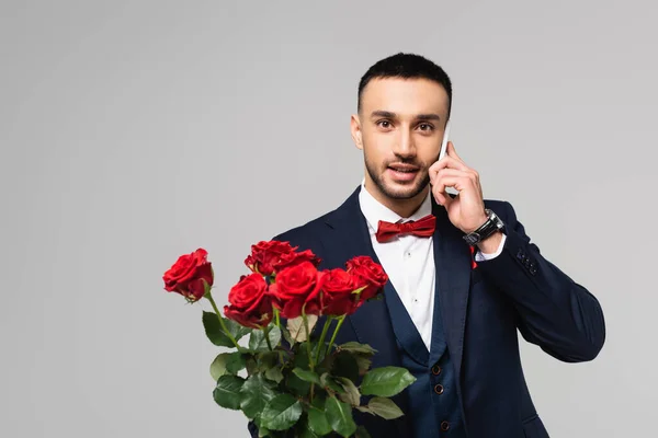 Morena hispano hombre con rosas rojas hablando en el teléfono móvil aislado en gris - foto de stock