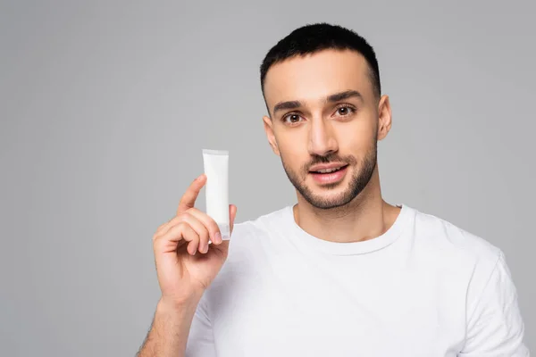 Giovane uomo ispanico che tiene il tubo di crema per le mani mentre guarda la fotocamera isolata sul grigio — Foto stock