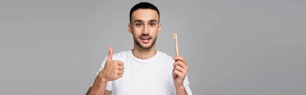 Alegre hombre hispano mostrando el pulgar hacia arriba mientras sostiene cepillo de dientes aislado en gris, bandera — Stock Photo