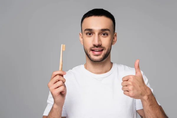 Hombre hispano positivo en camiseta blanca sosteniendo cepillo de dientes y mostrando el pulgar hacia arriba aislado en gris - foto de stock