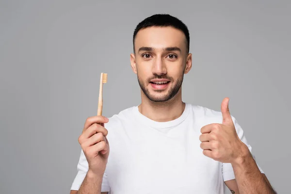 Positivo ispanico uomo in bianco t-shirt tenendo spazzolino da denti e mostrando pollice in alto isolato su grigio — Foto stock