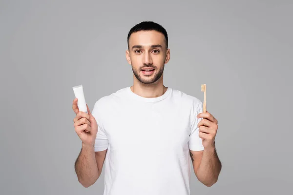 Hombre hispano positivo en camiseta blanca sosteniendo pasta de dientes y cepillo de dientes aislados en gris - foto de stock