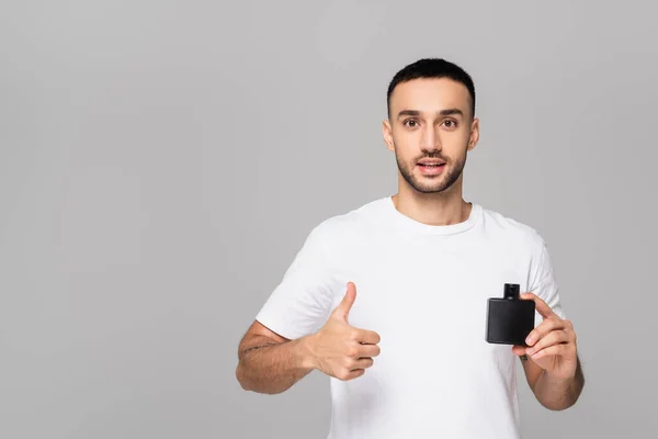 Positiver hispanischer Mann zeigt Daumen nach oben, während er Eau de Cologne isoliert auf grau hält — Stockfoto