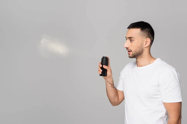 Bearded hispanic man in white t-shirt spraying deodorant isolated on grey — Stock Photo