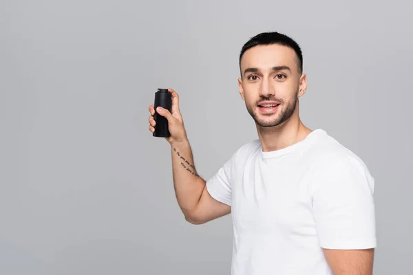 Morena hombre hispano en camiseta blanca mirando a la cámara mientras sostiene desodorante aislado en gris - foto de stock