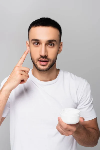 Молодий іспаномовний чоловік вказує на застосування крему для обличчя ізольовано на сірому — стокове фото