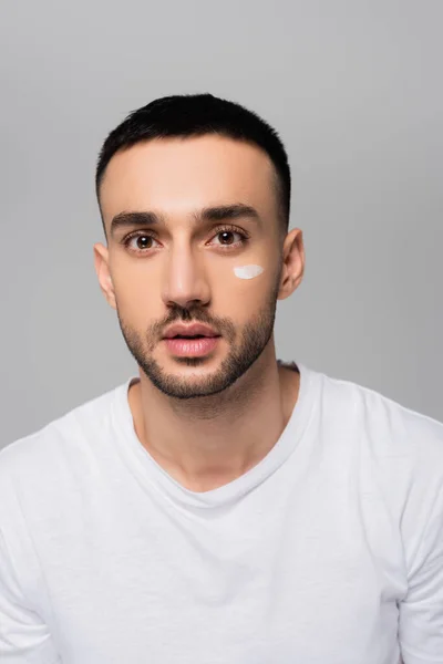 Young hispanic man with stroke of cosmetic cream on face looking at camera isolated on grey — Stock Photo