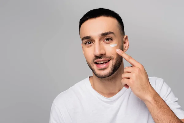 Fröhlicher hispanischer Mann, der Gesichtscreme isoliert auf grau aufträgt — Stockfoto