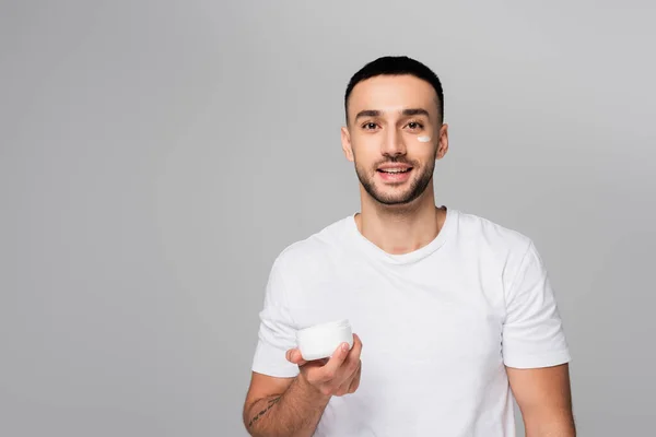 Morena hispânico homem sorrindo para a câmera enquanto segurando rosto creme isolado no cinza — Fotografia de Stock