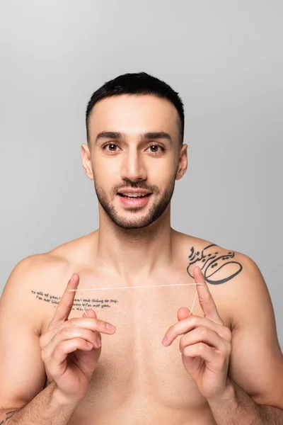 Smiling, tattooed hispanic man looking at camera while holding dental floss isolated on grey — Stock Photo