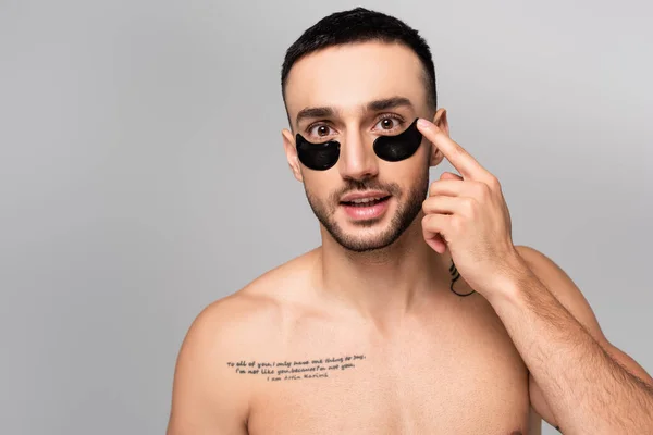 Латиноамериканец без рубашки, накладывающий повязки на глаза, глядя на камеру, изолированную на сером — стоковое фото