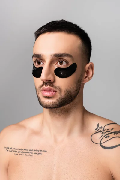 Hombre hispano tatuado y sin camisa con parches en los ojos mirando hacia otro lado aislado en gris - foto de stock