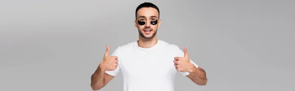 Uomo ispanico sorridente con macchie sugli occhi che mostrano i pollici in alto isolato sul grigio, banner — Foto stock