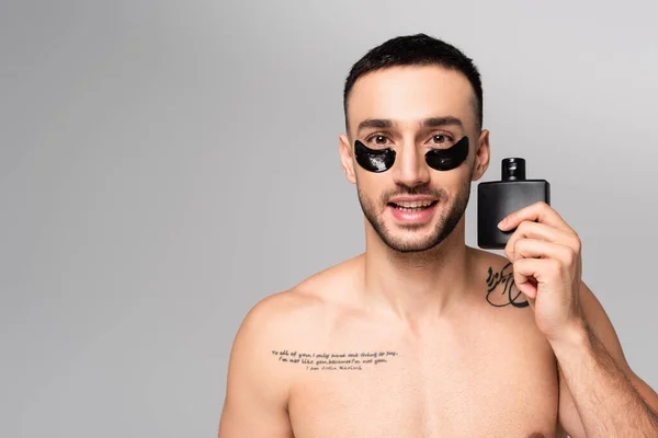 Felice, uomo ispanico a torso nudo guardando la macchina fotografica mentre tiene eau de cologne isolato sul grigio — Foto stock