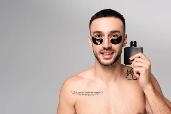 Feliz, hombre hispano sin camisa mirando a la cámara mientras sostiene a Eau de Cologne aislado en gris - foto de stock