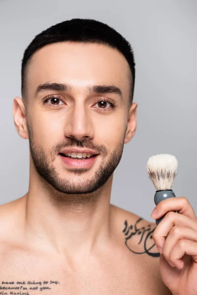 Homme hispanique tatoué souriant à la caméra tout en tenant la brosse à raser isolé sur gris — Photo de stock