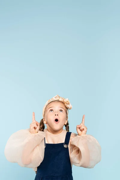 Schockiertes Kind im Stirnband mit Fliege, die mit den Fingern zeigt, während es isoliert auf blau blickt — Stockfoto