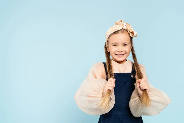 Cheerful kid touching pigtails and smiling isolated on blue — Stock Photo