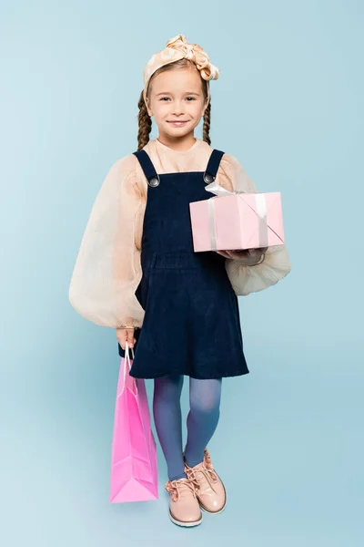 Full length of happy kid with pigtails holding shopping bag and present on blue — Stock Photo
