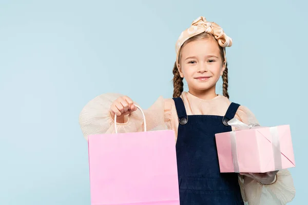 Criança feliz com tranças segurando saco de compras e presente isolado em azul — Fotografia de Stock